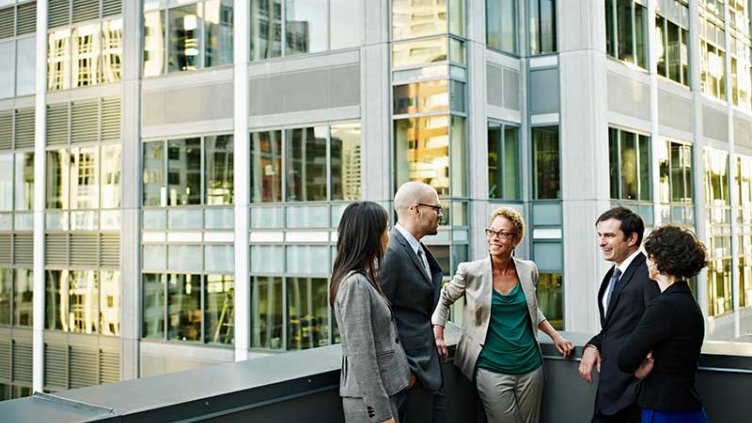 Group of coworkers standing in discussion on deck of office buildings in background