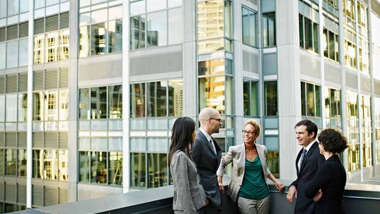 Group of coworkers standing in discussion on deck of office buildings in background