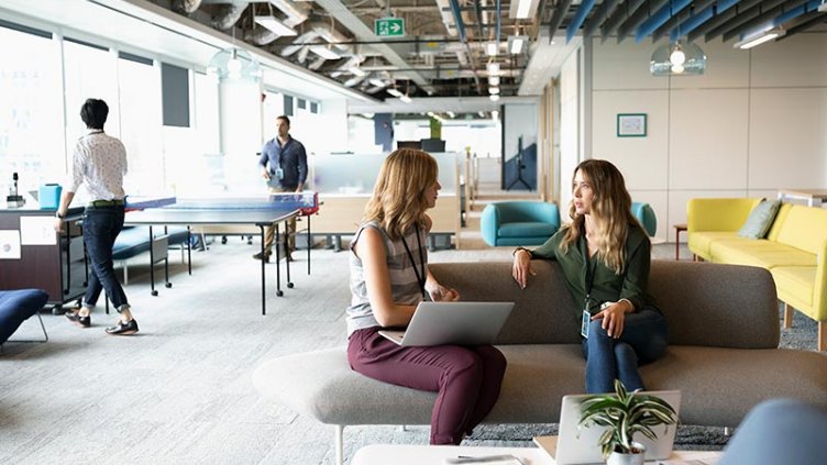 Creative businesswomen with laptop talking in open plan office with businessmen playing ping pong in background