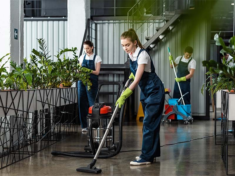 young cleaner vacuuming floor in office near colleagues