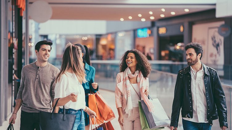 A group of boys and girls shopping together inside a bustling shopping center. They're browsing stores, carrying bags, and chatting