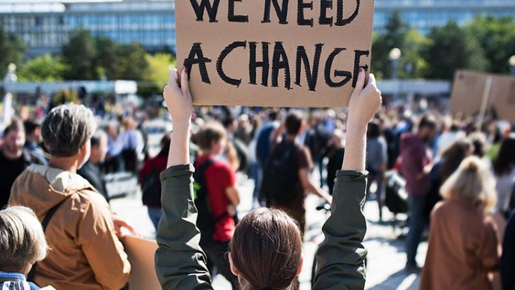 A lady standing in crowd by holding a bord of need a change