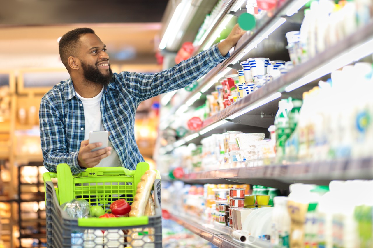 Male Buyer Shopping Groceries In Supermarket Taking Dairy Product From Shelf Standing With Shop Cart Indoors. Guy Buys Grocery Choosing Food In Super Market. Empty Space