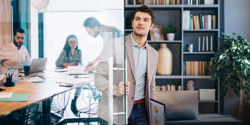 Man walking into room holding papers while people work at desks behind him